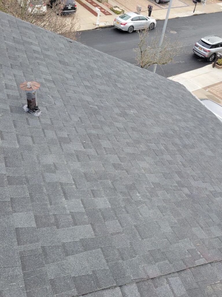 Aerial view of a gray asphalt shingle roof with a rusted vent pipe and flashing, overlooking a street with parked cars and sidewalks.