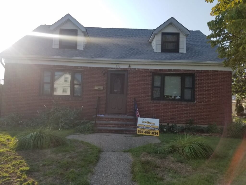 A brick Cape Cod style home with two front dormers and a newly installed gray shingle roof, with a yellow roofing contractor yard sign and small American flag near the front steps.
