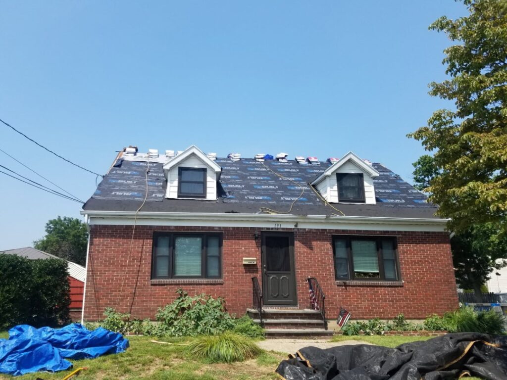 A red brick Cape Cod style home mid-roof-replacement, with synthetic underlayment covering the roof deck, shingle bundles staged along the ridge, two dormer windows, and blue and black tarps spread across the front lawn under a clear blue sky.