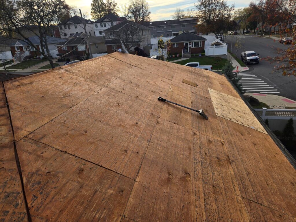 Aerial view of a residential roof stripped down to bare plywood sheathing, with a roofing shovel resting on the deck and a suburban street with homes, parked trucks, and autumn trees in the background.