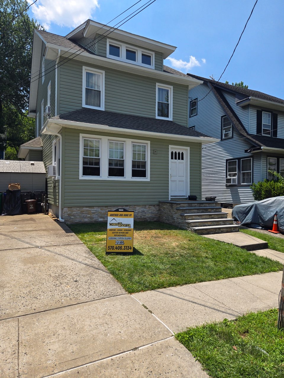 Two-story Staten Island home with sage green vinyl siding, stone veneer skirting and a Chris the Roofer yard sign on the lawn